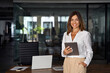 © Stock 4 You - Latin Hispanic mature adult professional business woman looking at camera and smiling. European businesswoman CEO holding digital tablet using fintech tab application standing at workplace in office.