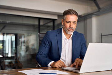 Mature Indian or Latin business man ceo trader using computer, typing, working in modern office, doing online data market analysis, thinking planning tech strategy looking at laptop with copy space.