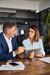 © Stock 4 You - Mature 50s age Latin business man mentoring mid age European business woman discussing project on tablet in office. Two colleagues, group of partners, professional business people working together.