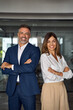 © Stock 4 You - Vertical portrait of smiling mature Latin hispanic business man and European business woman standing arms crossed in office. Diverse colleagues, group team of confident professional business people.