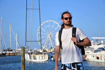 Young caucasian man with long hair standing on a port pier wearing casual beach clothing and a bagpack on a sunny summer day. 