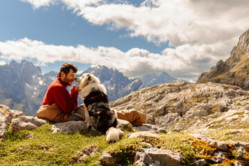 Bearded white man, mountaineer, having coffee for breakfast inside a sleeping bag after bivouacing in the mountains with his border collie dog. Traveling with a pet. Sport and adventure.