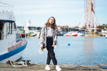 Beautiful Girl Standing Near Yacht On Pier. Seaport Or Berth In Background. Young Woman Wearing Casual Outfit Posing Near Water In Sunny Day. People Lifestyle And Rest Concept. Blurred Background