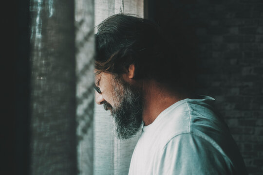 One lonely and depresse man with eyes closed stand up against the window at home. People and stress or desperation. Mature male with beard sad alone indoor. Concept of unhappy person lifestyle. Mental