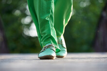 A Woman In Green Trousers And Moccasins Walks Along The Road In Summer