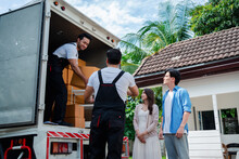 Asian Couple Check While Unloading Boxes And Furniture From A Pickup Truck To A New House With Service Cargo Two Men Movers Worker In Uniform Lifting Boxes. Concept Of Home Moving And Delivery.