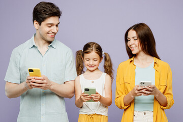 Wall Mural - Young happy parents mom dad with child kid daughter girl 6 years old wear blue yellow casual clothes hold in hand use look at mobile cell phone isolated on plain purple background. Family day concept.