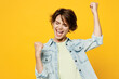 © ViDi Studio - Young happy fun woman she wear green t-shirt denim shirt casual clothes doing winner gesture celebrate clenching fists say yes isolated on plain yellow background studio portrait. Lifestyle concept.
