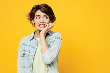 Wall Mural - Young minded pensive dissatisfied sad woman she wears green t-shirt denim shirt casual clothes look aside biting nails fingers isolated on plain yellow background studio portrait. Lifestyle concept.