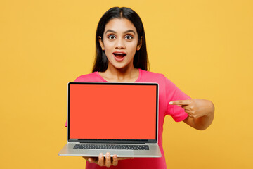 Young surprised IT Indian woman wearing pink t-shirt casual clothes hold use work point on laptop pc computer with blank screen workspace area isolated on plain yellow background. Lifestyle concept.