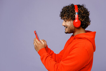 Wall Mural - Side view young Indian man wear red orange hoody casual clothes listen to music in headphones use mobile cell phone isolated on plain pastel purple color background studio portrait. Lifestyle concept.