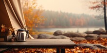 Wooden Table With A Kettle In Front Of A Blurred Background Of Autumn Nature On A Campsite. Lake, Stones And Forest In Cloudy Weather. For Product Display.