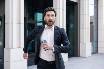 A young man businessman in a business suit holds a mobile phone in his hands and runs on the street near the office center, is late for a meeting