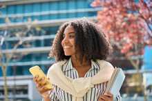 Smiling Happy Cute African Teen Girl Student Holding Cellphone Looking Away With Smartphone Technology Device In Hand Walking In College Park Outside Using Apps On Mobile Phone, Authentic Shot.