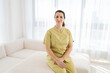 Portrait of smiling female masseuse therapist in uniform sitting posing on couch looking at camera with friendly expression, on background of window in modern wellness center. Concept of body care