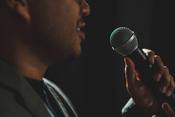 Canvas Print - Confident successful speaker man talking on stage with spotlight strike through the darkness at corporate business event. Public speaker giving talk at conference hall. Stand up comedian.