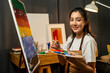 © Chanakon - Asian woman artist in the darkroom warm light working on painting with brush and variant acrylic color. Female artist painter on canvas in creative studio as art concept