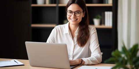 Poster - Beautiful smiling woman sitting at the table, using a laptop and writing in a notebook, taking notes.