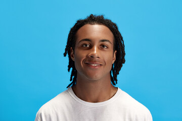 Wall Mural - Portrait of young african man, student with dreads in white t-shirt looking at camera and smiling against blue studio background. Concept of youth, human emotions, lifestyle, fashion, ad