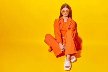 Wall Mural - Young fashion woman in orange pants orange top orange shirt on yellow background. Platform slides sandals, orange sunglasses.