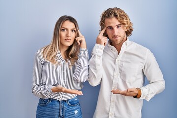 Canvas Print - Young couple standing over blue background confused and annoyed with open palm showing copy space and pointing finger to forehead. think about it.