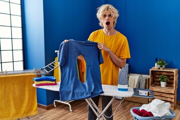 Sticker - Young blond man holding t shirt with burned hole with surprise expression at laundry room