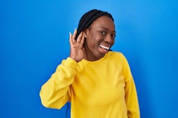 Wall Mural - Beautiful black woman standing over blue background smiling with hand over ear listening an hearing to rumor or gossip. deafness concept.