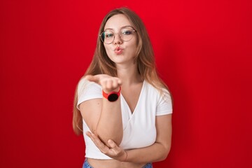 Sticker - Young caucasian woman standing over red background looking at the camera blowing a kiss with hand on air being lovely and sexy. love expression.