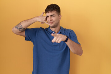 Sticker - Young hispanic man standing over yellow background smiling doing talking on the telephone gesture and pointing to you. call me.