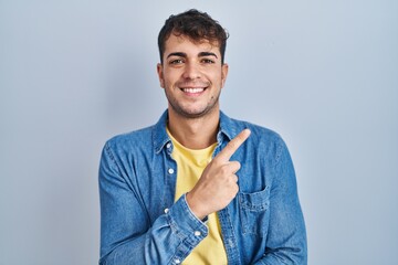 Wall Mural - Young hispanic man standing over blue background with a big smile on face, pointing with hand finger to the side looking at the camera.