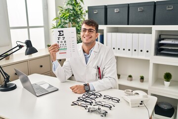 Sticker - Young hispanic man working at ophthalmology clinic looking positive and happy standing and smiling with a confident smile showing teeth