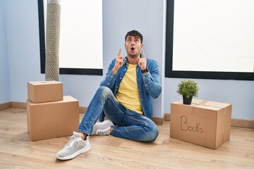 Wall Mural - Young hispanic man sitting on the floor at new home amazed and surprised looking up and pointing with fingers and raised arms.