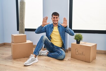 Wall Mural - Young hispanic man sitting on the floor at new home showing and pointing up with fingers number ten while smiling confident and happy.