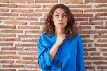 Canvas Print - Beautiful brunette woman standing over bricks wall thinking concentrated about doubt with finger on chin and looking up wondering