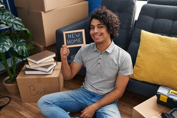 Poster - Hispanic man with curly hair sitting on the floor at new home looking positive and happy standing and smiling with a confident smile showing teeth