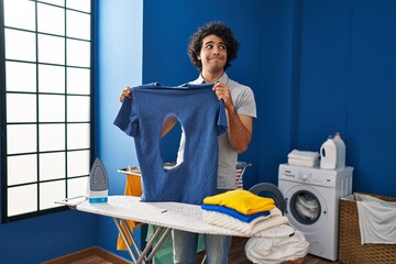 Wall Mural - Hispanic man with curly hair ironing holding burned iron shirt at laundry room smiling looking to the side and staring away thinking.