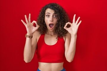 Sticker - Hispanic woman with curly hair standing over red background looking surprised and shocked doing ok approval symbol with fingers. crazy expression