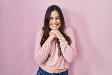 Canvas Print - Young brunette woman standing over pink background laughing nervous and excited with hands on chin looking to the side