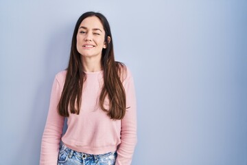 Poster - Young brunette woman standing over blue background winking looking at the camera with sexy expression, cheerful and happy face.