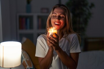 Wall Mural - Young blonde woman sitting on bed smelling aromatic candle at bedroom