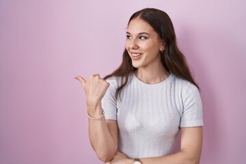 Wall Mural - Young hispanic girl standing over pink background smiling with happy face looking and pointing to the side with thumb up.