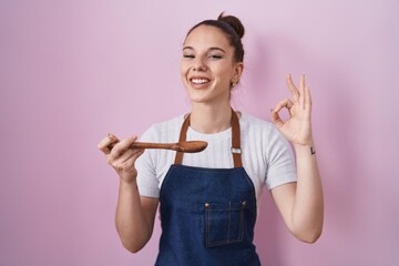 Poster - Young hispanic girl wearing professional cook apron holding wood spoon smiling and laughing hard out loud because funny crazy joke.