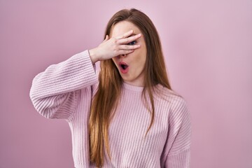 Wall Mural - Young caucasian woman standing over pink background peeking in shock covering face and eyes with hand, looking through fingers with embarrassed expression.