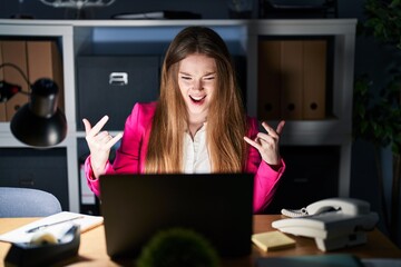 Canvas Print - Young caucasian woman working at the office at night shouting with crazy expression doing rock symbol with hands up. music star. heavy concept.