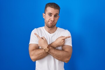 Poster - Young caucasian man standing over blue background pointing to both sides with fingers, different direction disagree