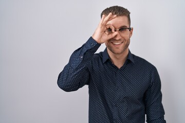 Sticker - Young caucasian man standing over isolated background doing ok gesture with hand smiling, eye looking through fingers with happy face.