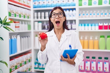 Poster - Young hispanic woman working at pharmacy drugstore holding red heart angry and mad screaming frustrated and furious, shouting with anger looking up.