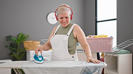 Wall Mural - Middle age grey-haired woman listening to music ironing clothes at laundry room