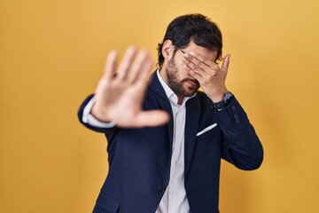 Sticker - Handsome latin man standing over yellow background covering eyes with hands and doing stop gesture with sad and fear expression. embarrassed and negative concept.