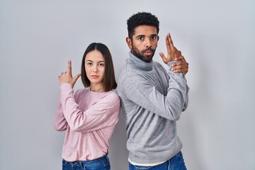 Poster - Young hispanic couple standing together holding symbolic gun with hand gesture, playing killing shooting weapons, angry face